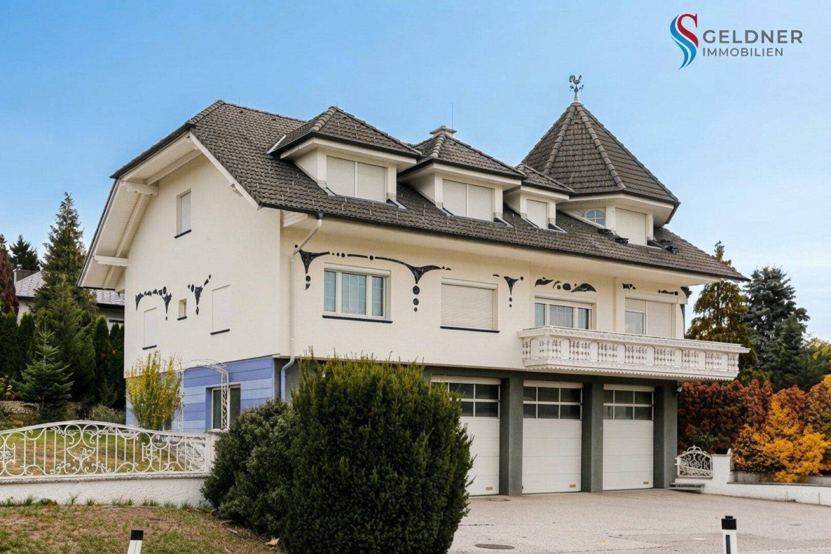 Ansicht des Hauses von der Seite mit Turmdach, Balkon und Garagen im Untergeschoss, umgeben von Bäumen.