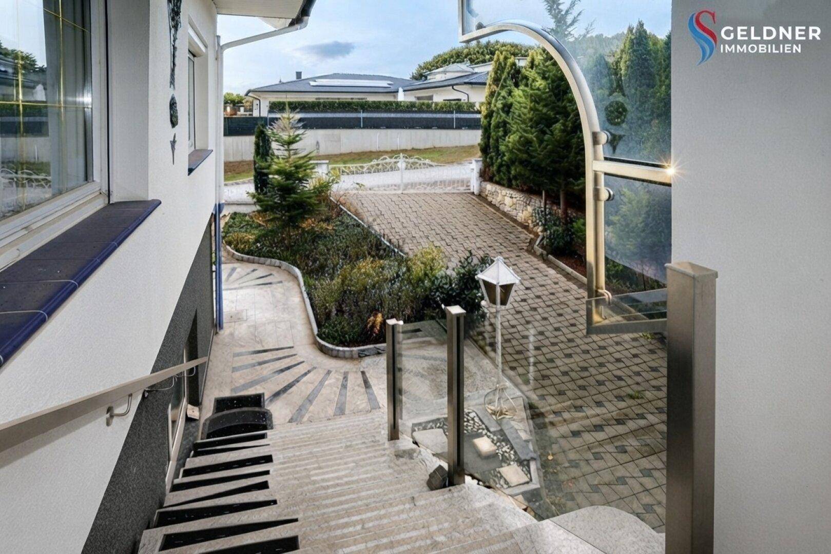 Eingangsbereich mit gepflastertem Weg, Treppe und moderner Glasüberdachung, umgeben von Grün.