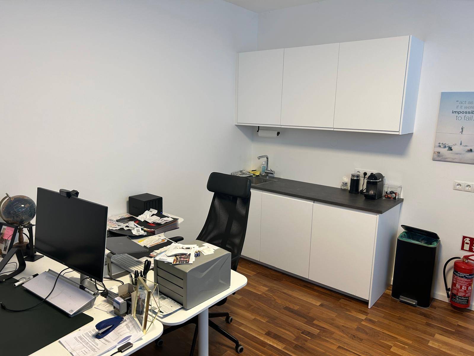 Kleine Büroküche mit weißen Hängeschränken, Spülbecken und Arbeitsplatte, integriert in den Arbeitsbereich.