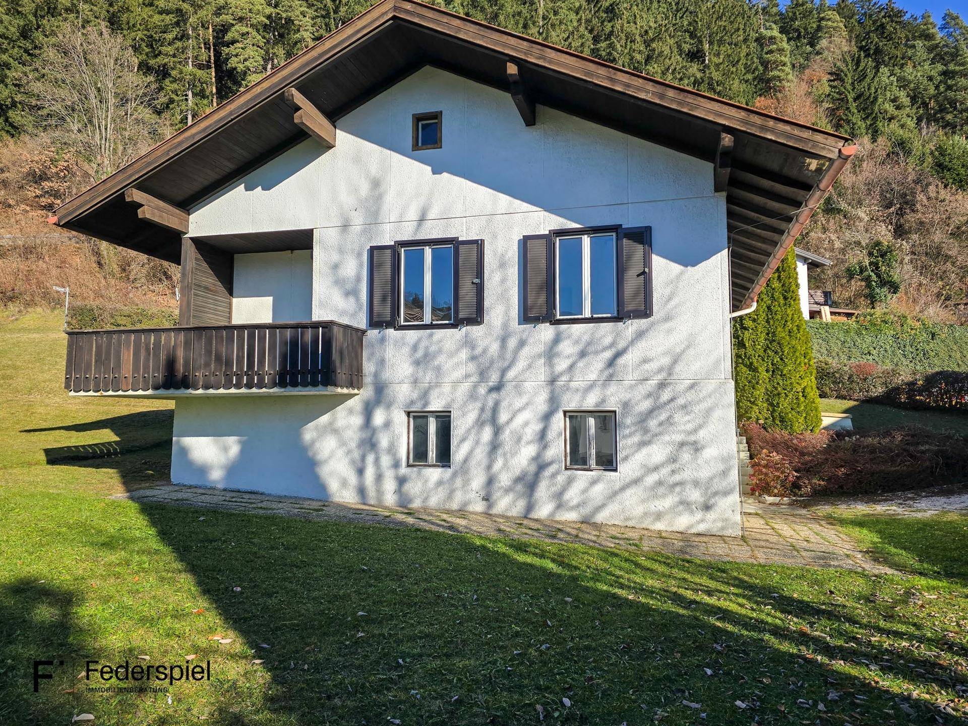 Seitenansicht des Hauses mit Balkon und Blick auf den bewaldeten Hang unter blauem Himmel.