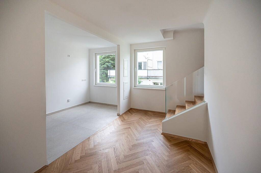 Helles Zimmer mit Fischgrätparkett und Zugang zu einem weiteren Bereich sowie einer modernen Treppe.