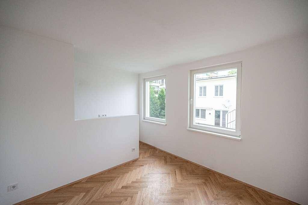 Unmöbliertes Zimmer mit Fischgrätparkett und zwei Fenstern, die viel Tageslicht hereinlassen.