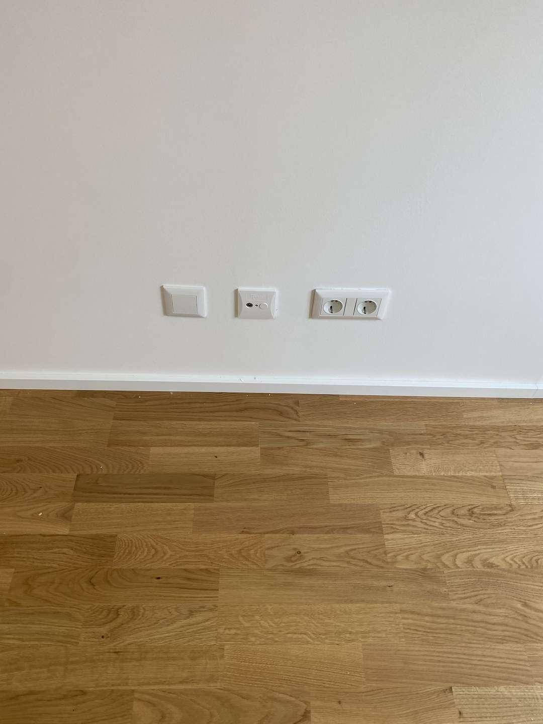 Detailaufnahme von Steckdosen und Schaltern an einer weißen Wand über dem Parkettboden.