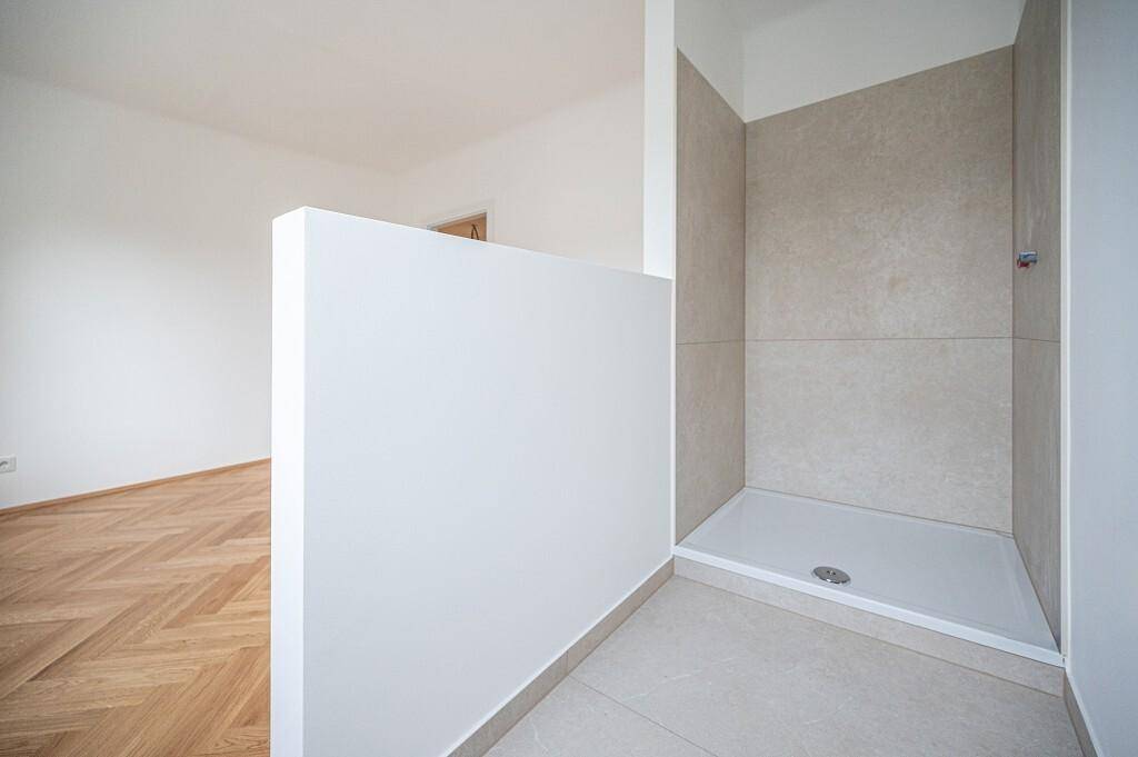 Modernes Badezimmer mit begehbarer Dusche und hellen Fliesen, angrenzend an einen Raum mit Parkett.