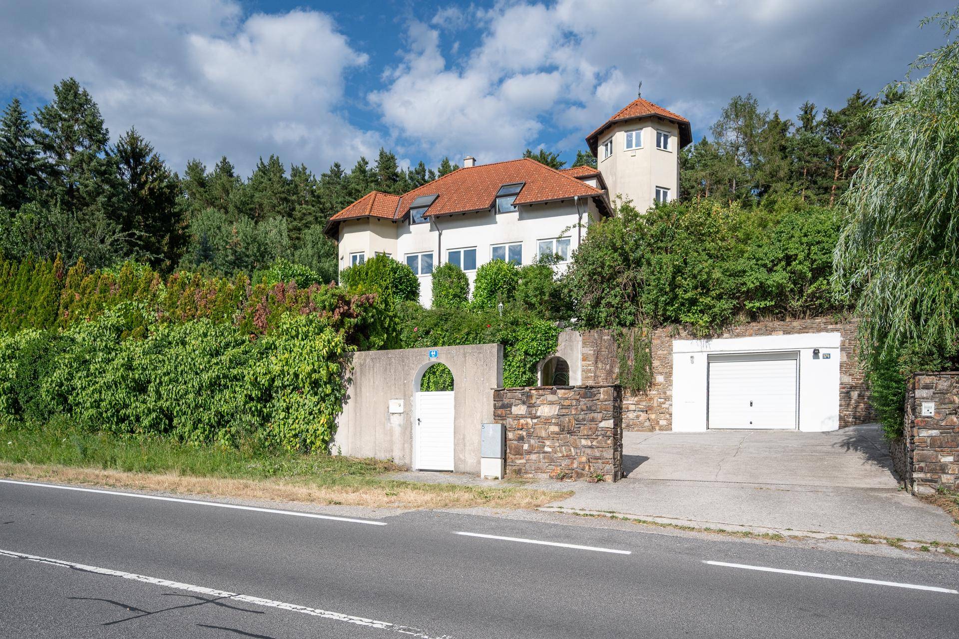 Ansicht des Hauses von der Straße mit Turm, Garage und üppiger Bepflanzung.
