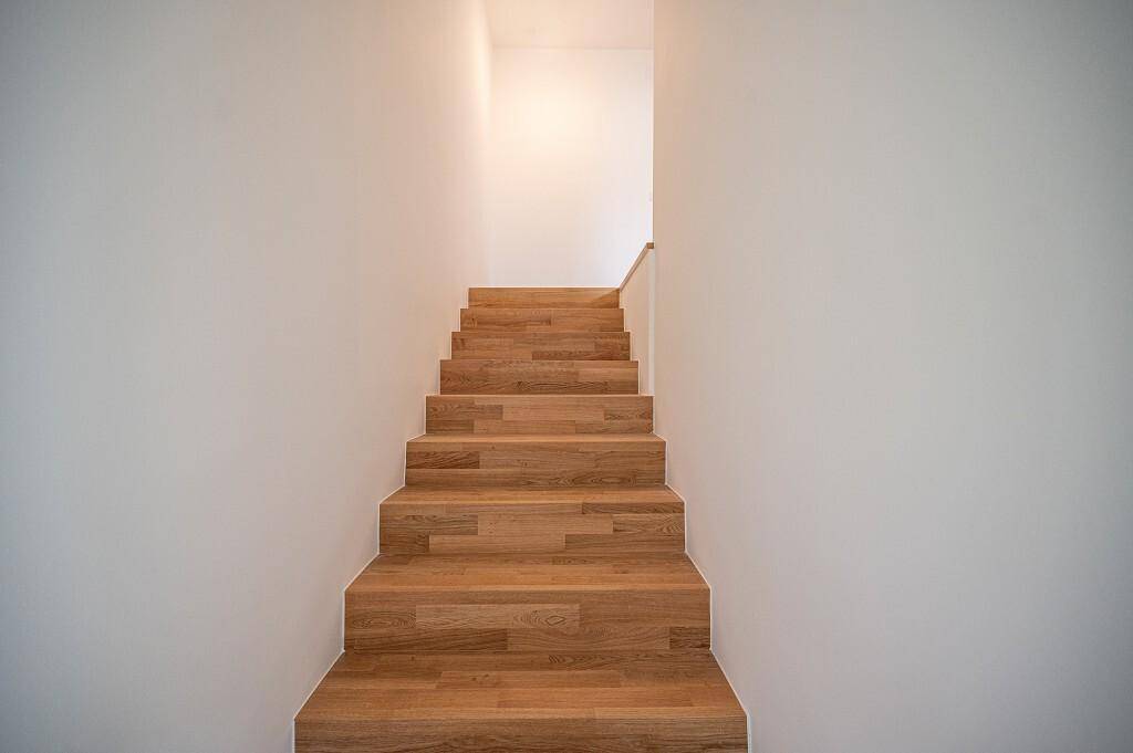 Moderne Holztreppe mit weiß gestrichenen Wänden, die in ein helles Obergeschoss führt.