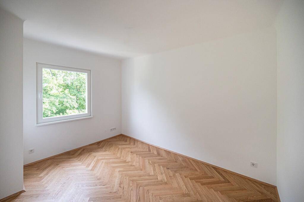 Helles Schlafzimmer mit Fischgrätparkett und einem Fenster mit Blick ins Grüne.