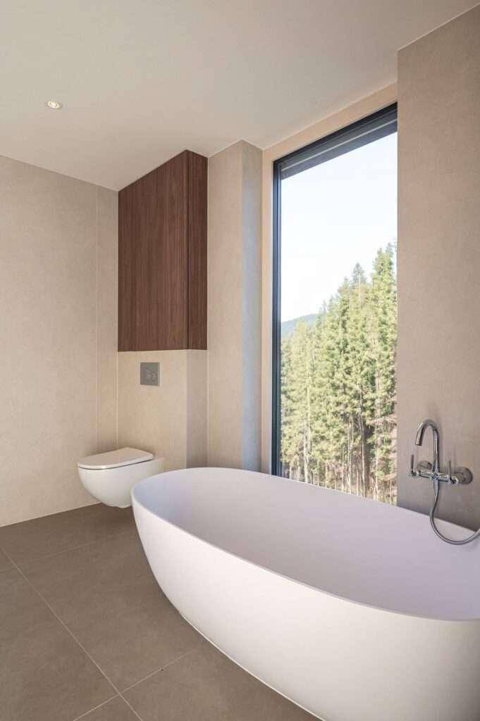 Luxuriöses Badezimmer mit freistehender Badewanne, wandhängender Toilette und großem Fenster mit Waldblick.