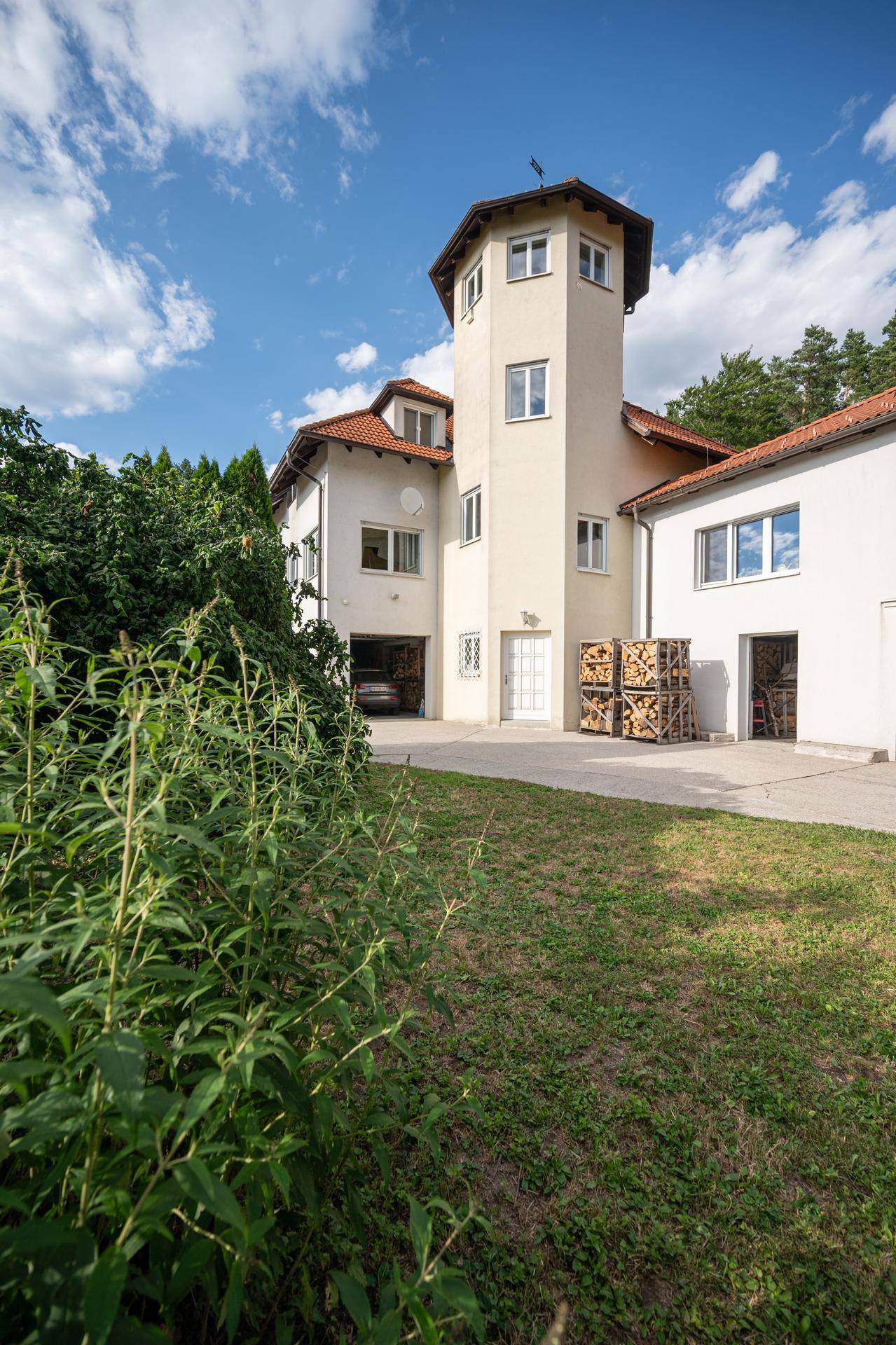 Seitenansicht des Hauses mit markantem Turm, Rasenfläche und Holzstapeln.