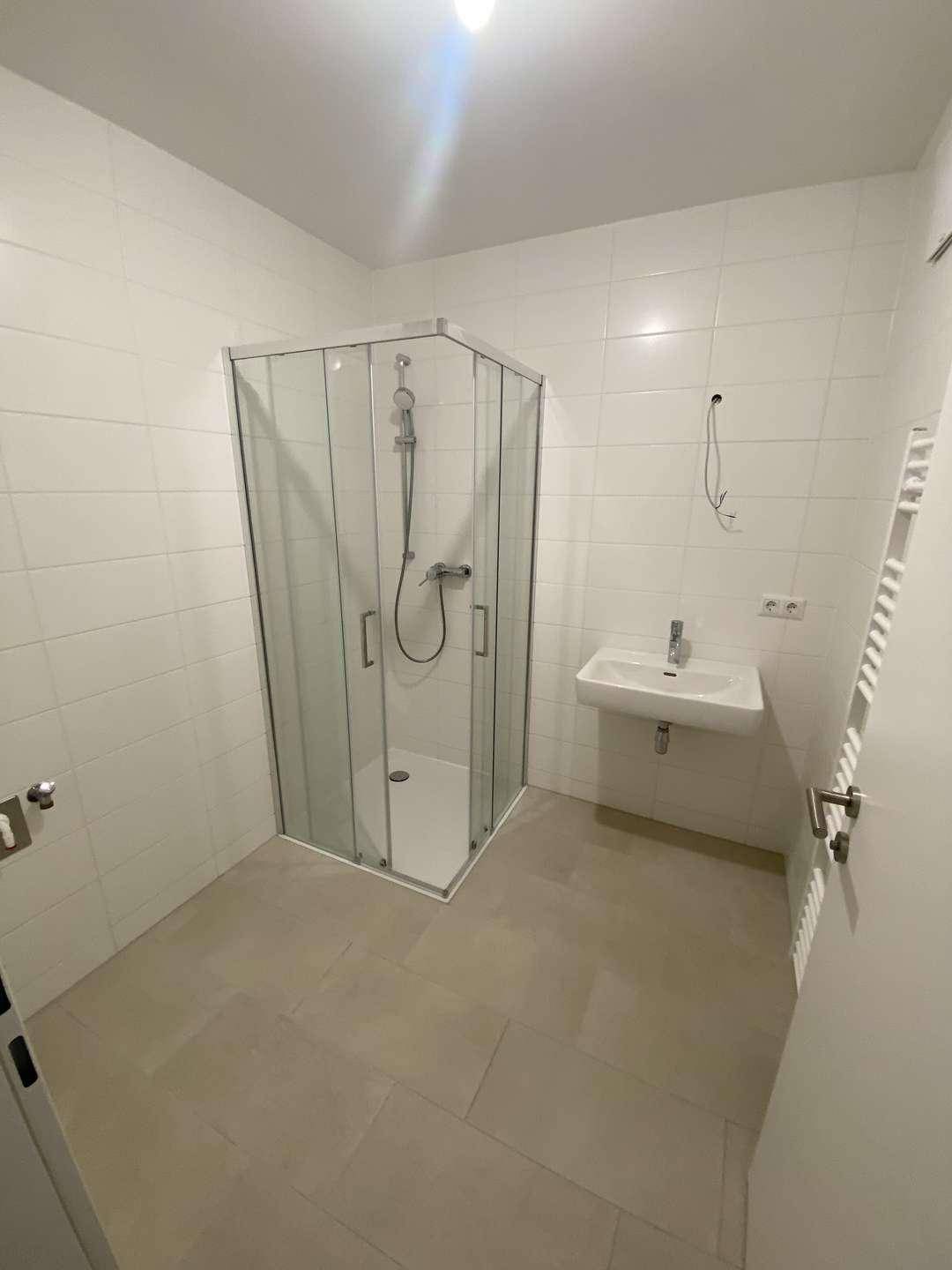 Helles Badezimmer mit Duschkabine, Waschbecken und weißen Wandfliesen sowie einem Handtuchheizkörper.
