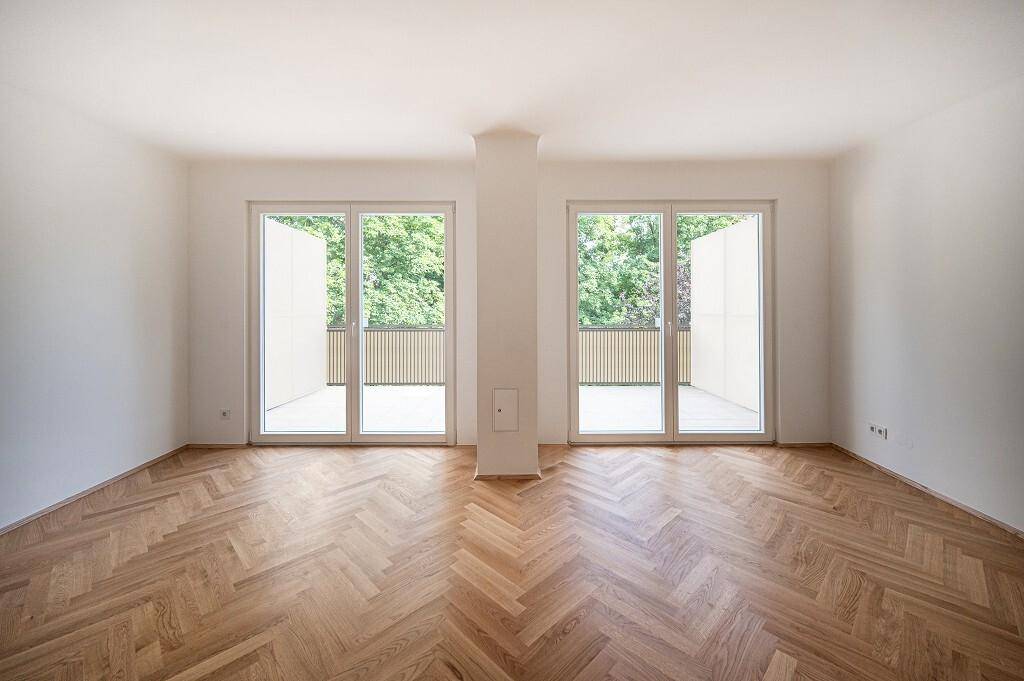 Geräumiges Wohnzimmer mit Fischgrätparkett und zwei großen Fenstertüren, die zum Balkon führen.
