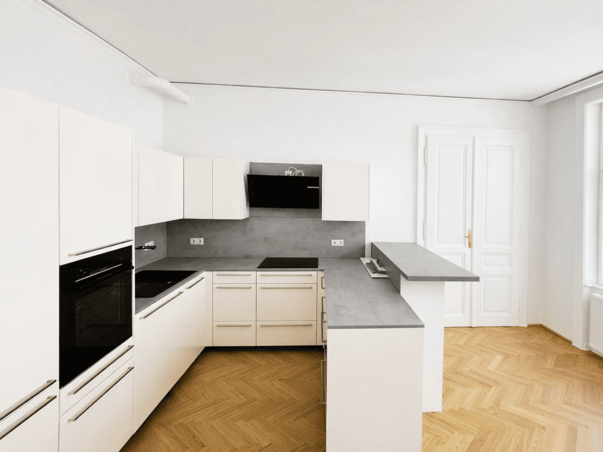 Moderne Einbauküche in Weiß mit grauer Arbeitsplatte und Fischgrätparkettboden.