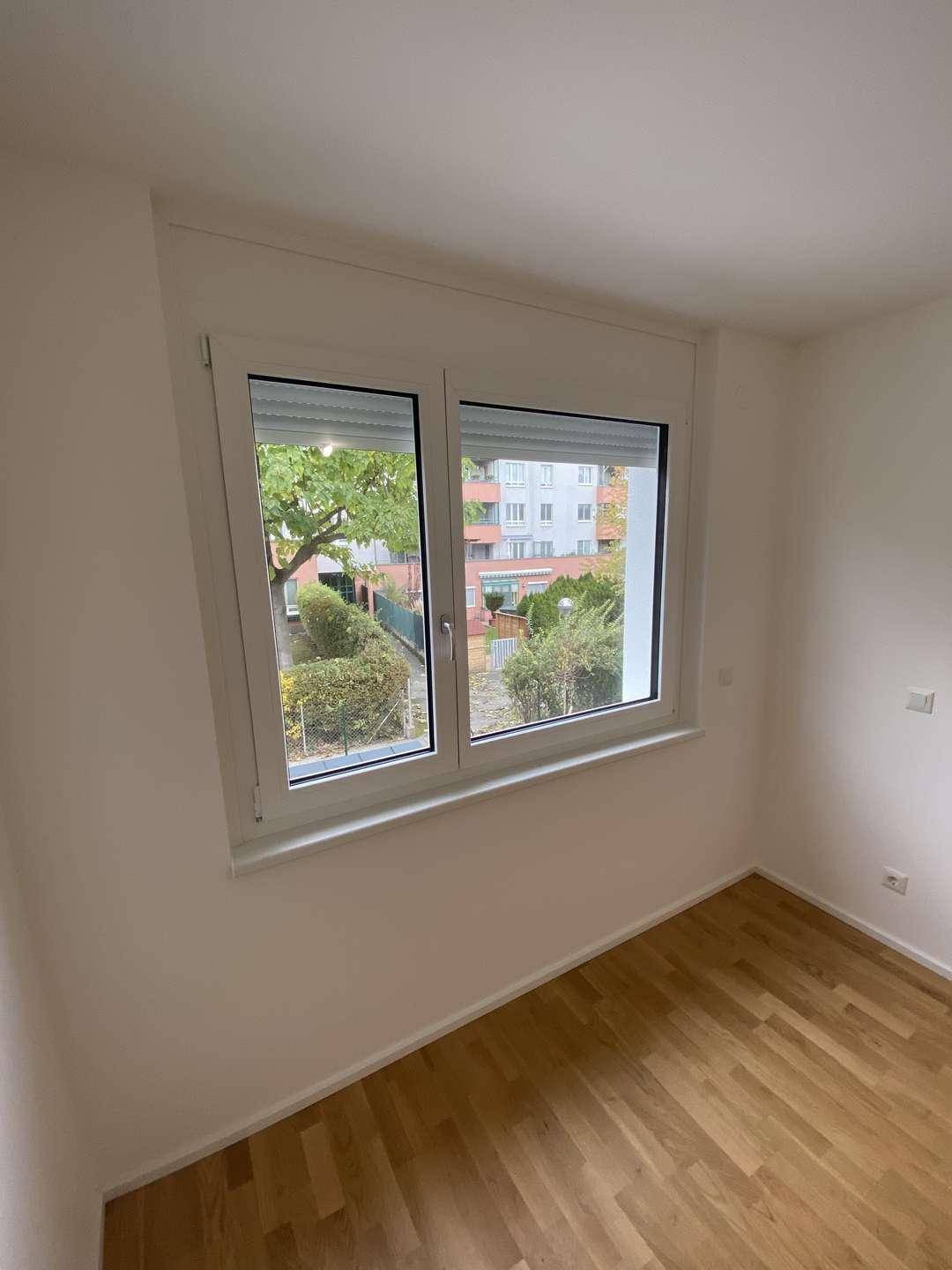 Helles Zimmer mit Parkettboden und Fenster mit Blick auf die grüne Umgebung und Rollläden.