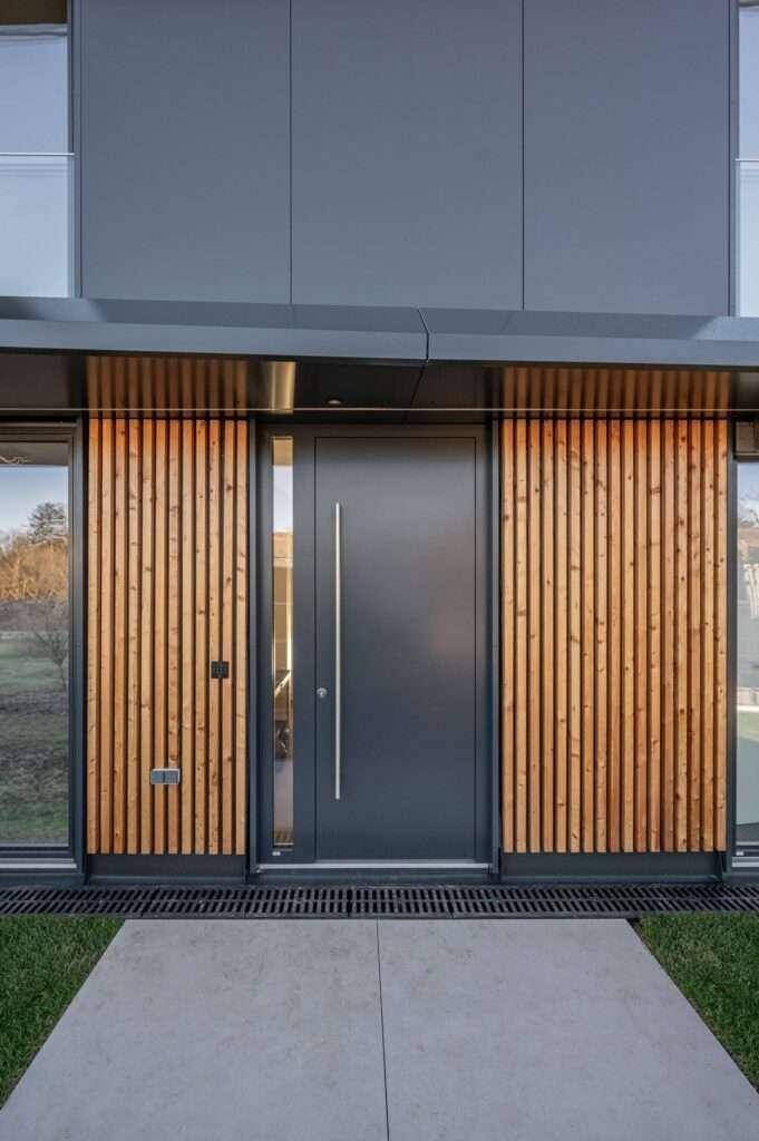 Moderner Hauseingang mit dunkler Tür, vertikalen Holzlamellen und großen Glasflächen.