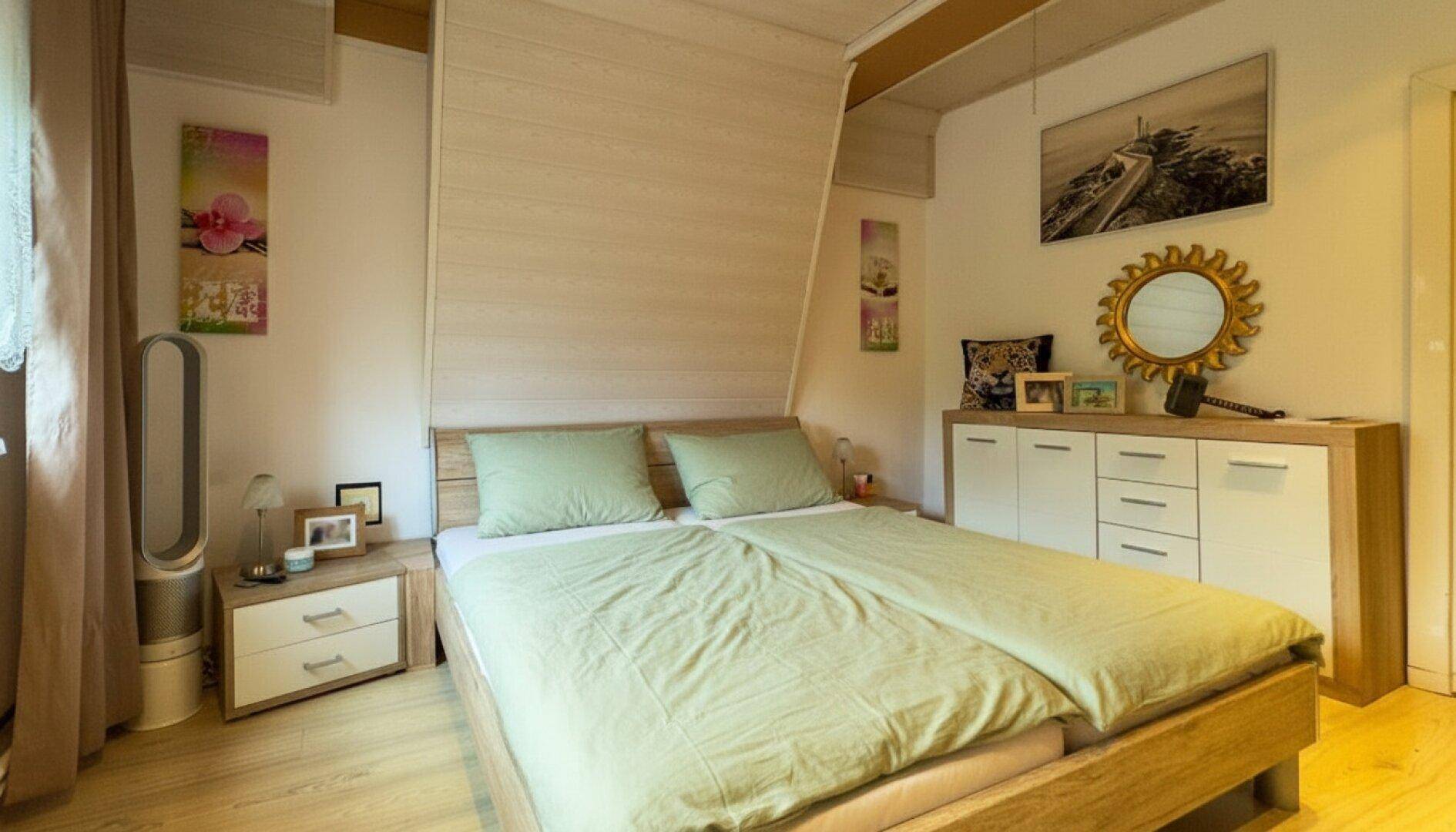 Helles Schlafzimmer mit Holzbett, Nachttischen und einer Kommode unter einer Dachschräge.