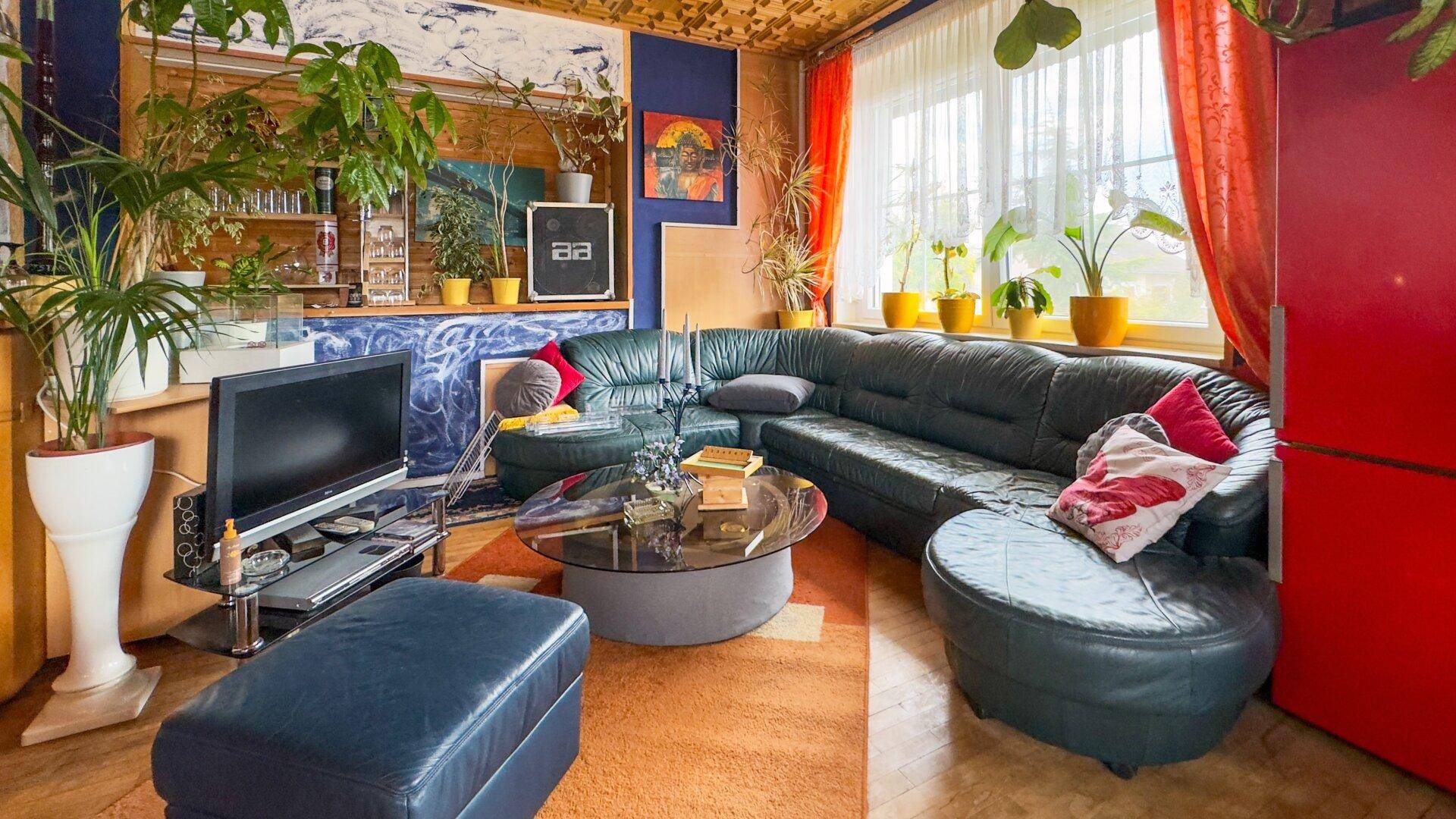 Farbenfrohes Wohnzimmer mit einer großen Ledercouch, Fernseher und zahlreichen Zimmerpflanzen, die eine lebendige Atmosphäre schaffen.