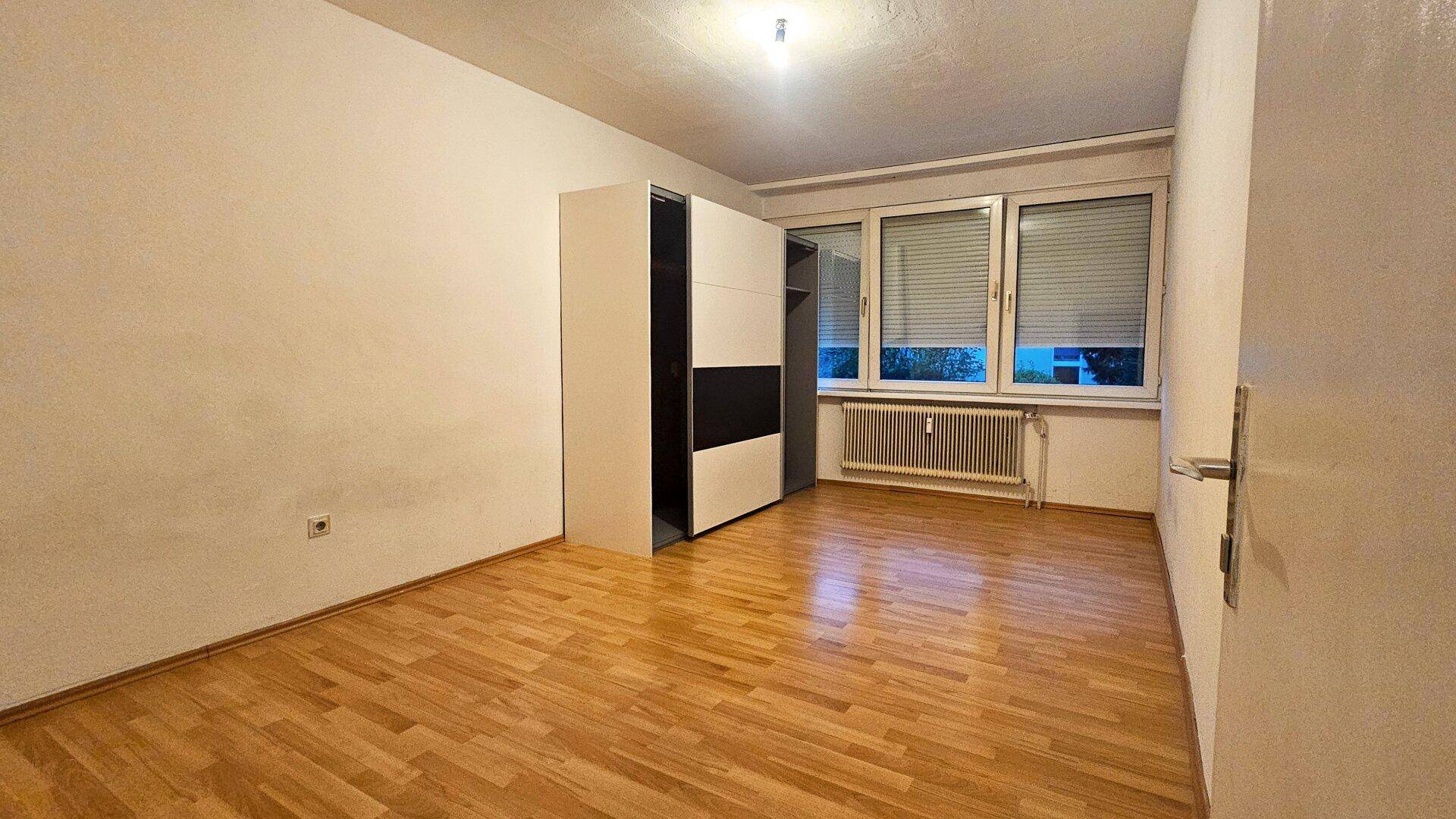 Helles Schlafzimmer mit Parkettboden, Fenster mit Jalousien und einem Kleiderschrank.