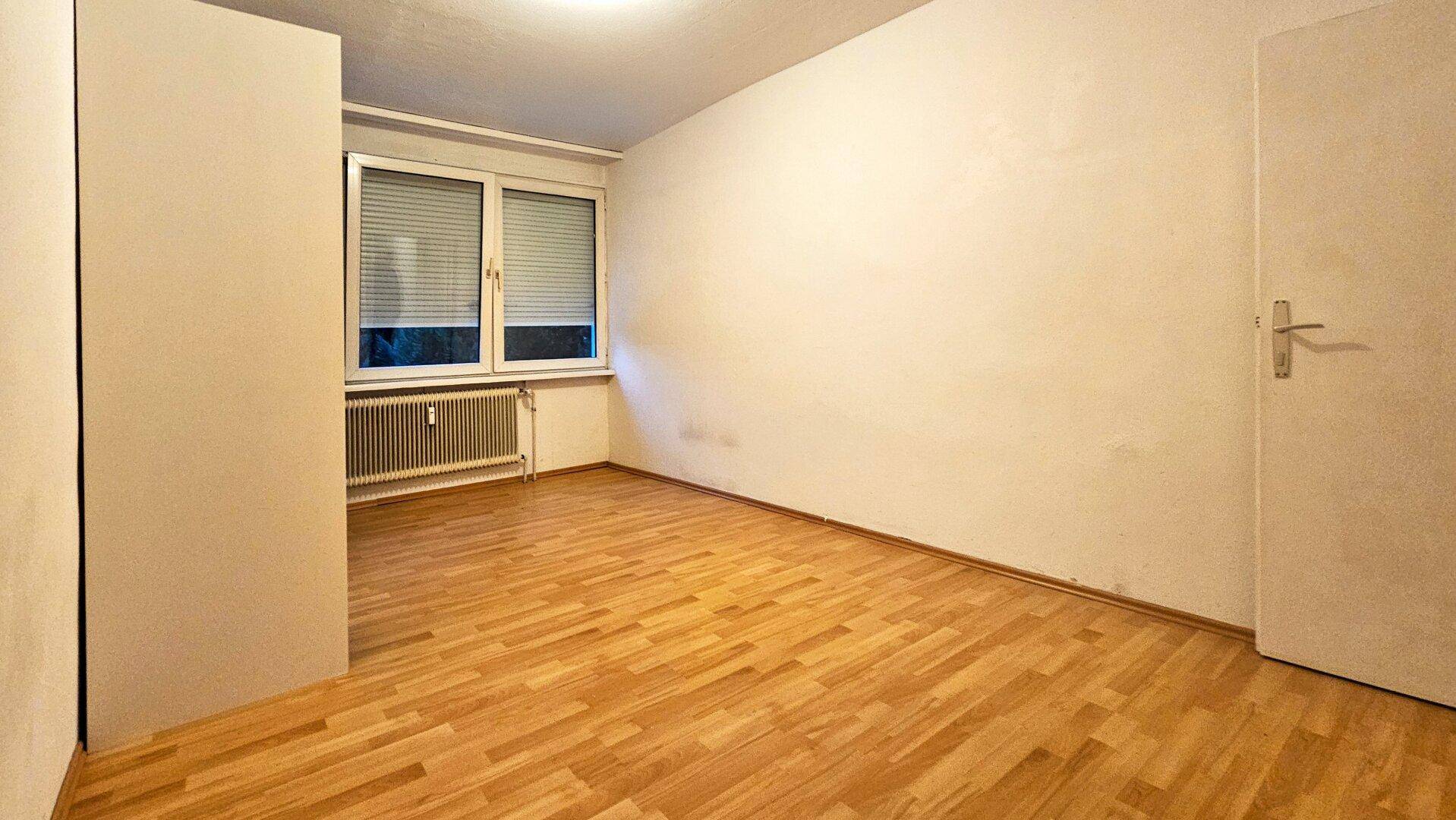 Leeres Schlafzimmer mit Parkettboden, Heizkörper unter dem Fenster und weiß gestrichenen Wänden.