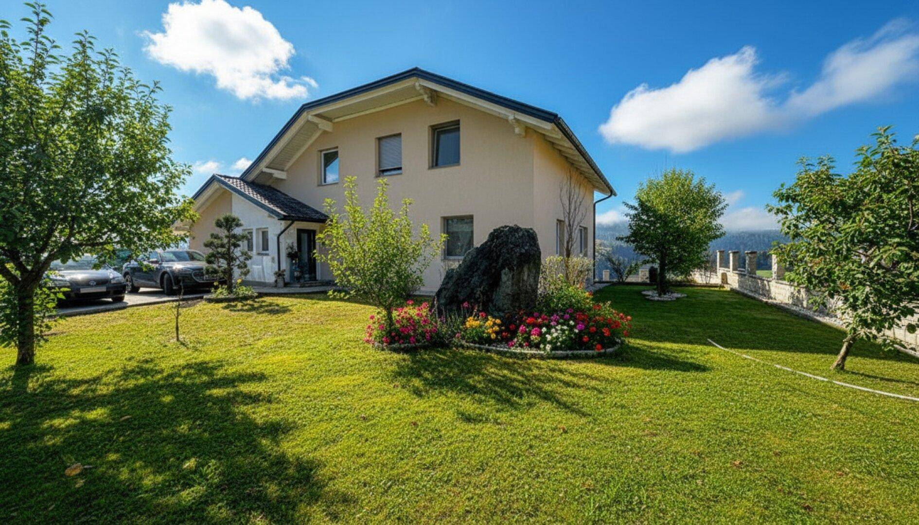 Gepflegter Garten mit großer Rasenfläche, Blumenbeeten und einem großen Felsen vor dem Haus.