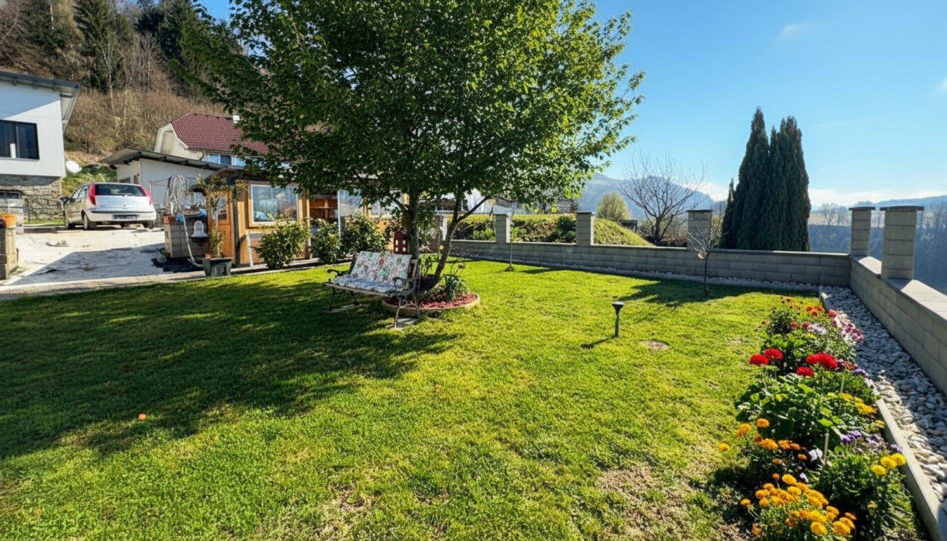Sonniger Garten mit Rasenfläche, Baum und einer Steinmauer, die einen weiten Blick bietet.