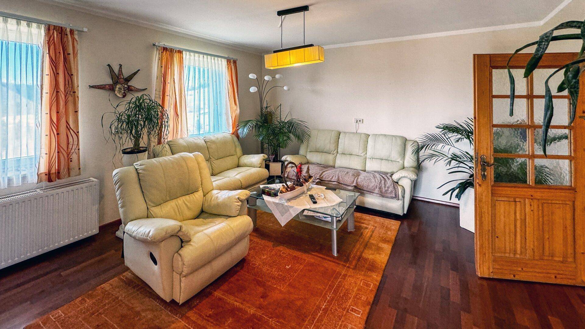 Gemütliches Wohnzimmer mit dunklem Holzboden, beigen Sofas und großen Fenstern.