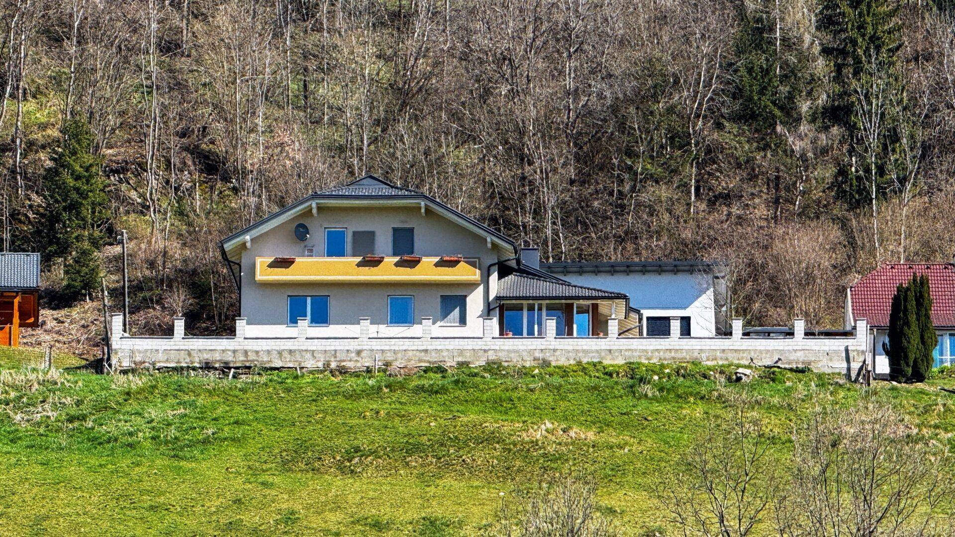 Haus in Hanglage mit grüner Umgebung und bewaldetem Hintergrund unter blauem Himmel.