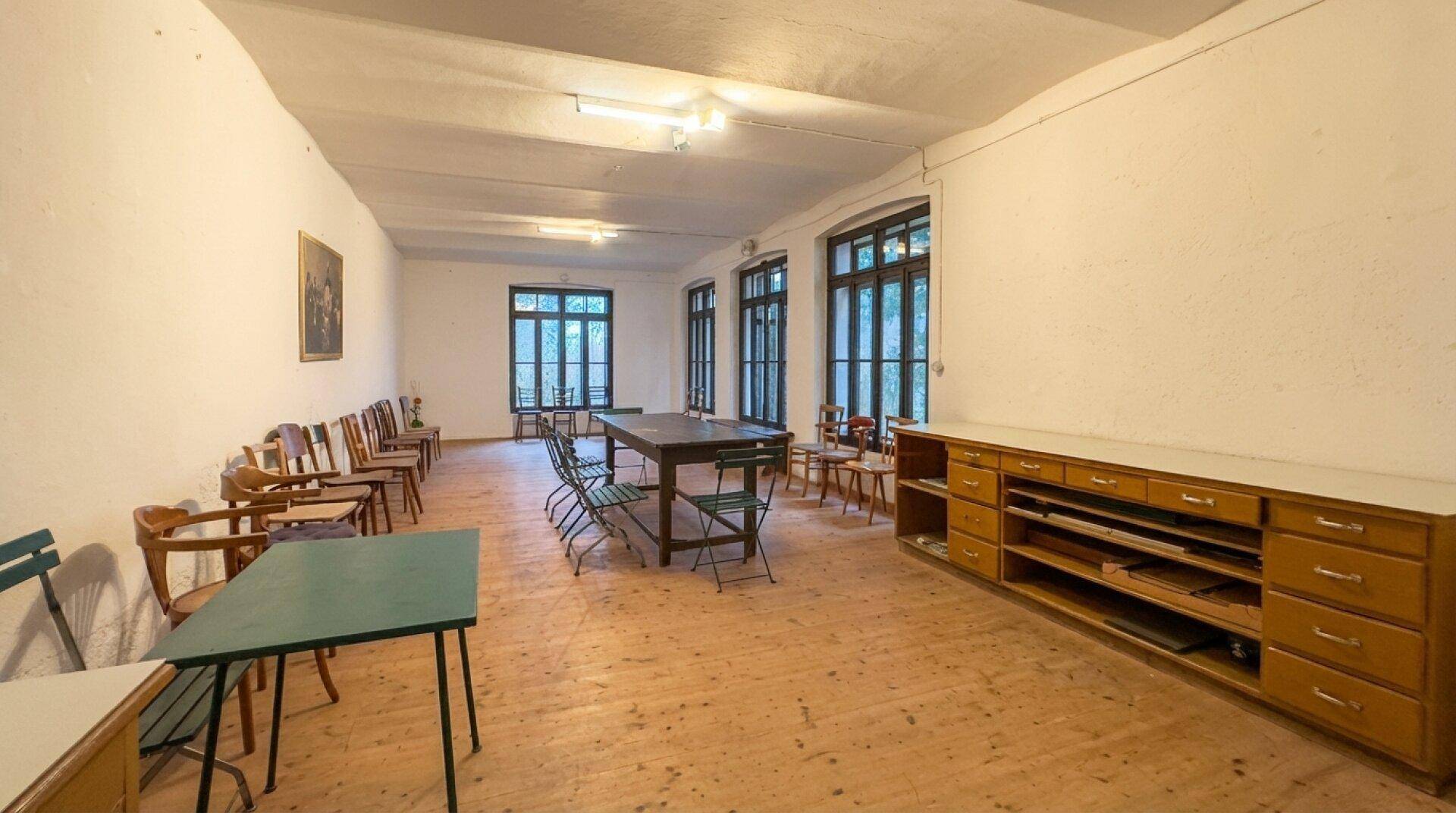 Großer Raum mit Holzboden, vielen Fenstern und einfacher Bestuhlung.