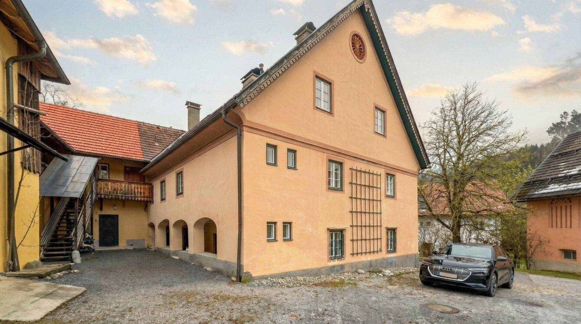 Historisches Gebäude mit Anbau, Holzbalkon und Kiesparkplatz, umgeben von Bäumen.