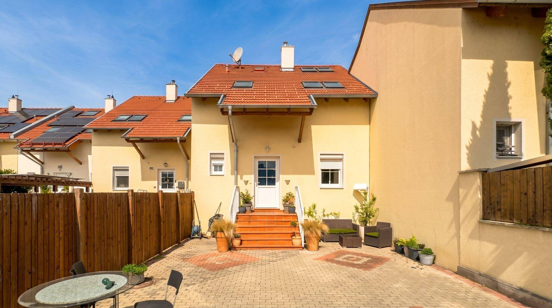 Sonniger Hinterhof mit gepflasterter Terrasse, Treppe zum Hauseingang und angrenzenden Gärten.