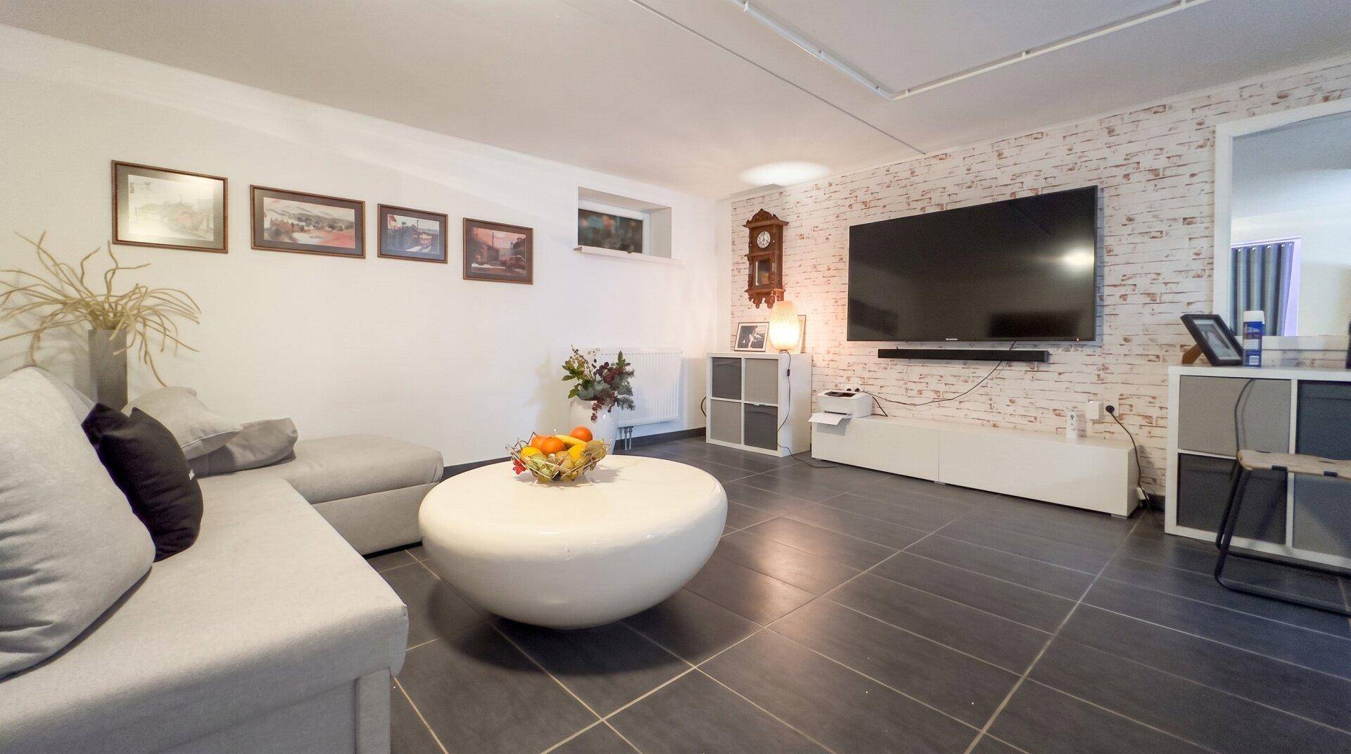Gemütlicher Wohnraum im Untergeschoss mit grauem Sofa, Ziegelwandakzent und Flachbildfernseher.
