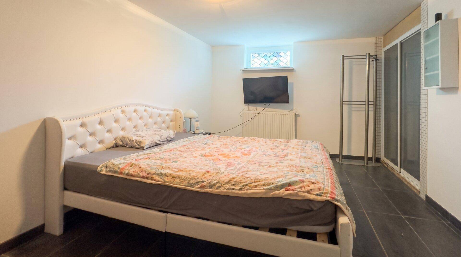Schlafzimmer im Untergeschoss mit gepolstertem Bett, kleinem Fenster und dunklen Bodenfliesen.