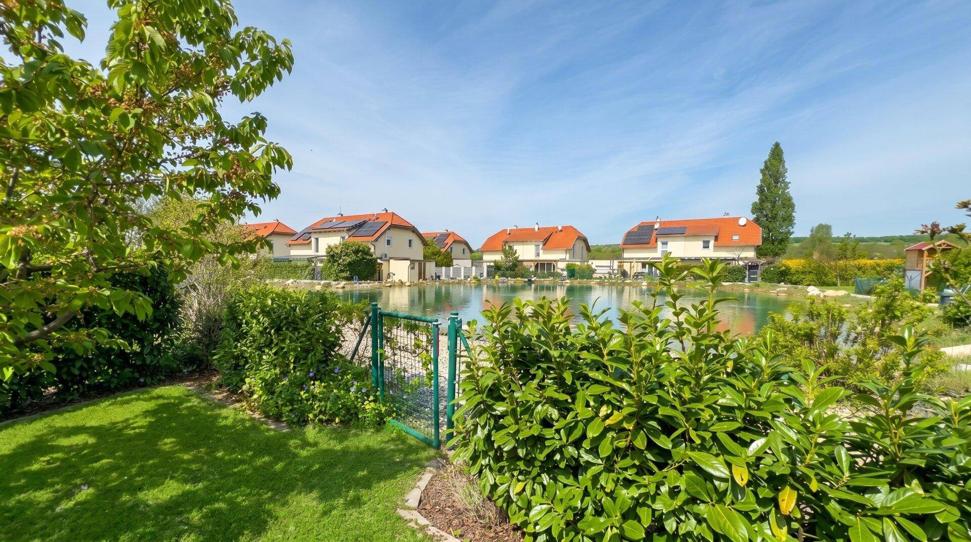 Malerischer Blick auf einen Teich mit umliegenden Häusern, umgeben von üppigem Grün.