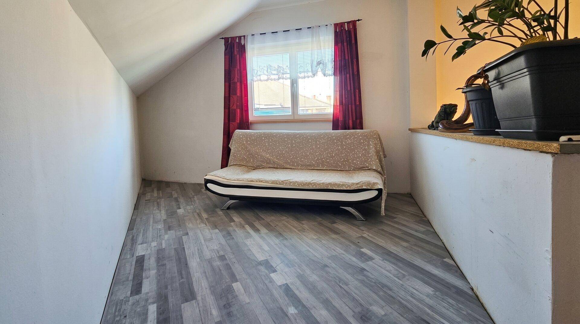 Dachzimmer mit schrägen Decken, Fenster mit roten Vorhängen und einem einfachen Schlafsofa.
