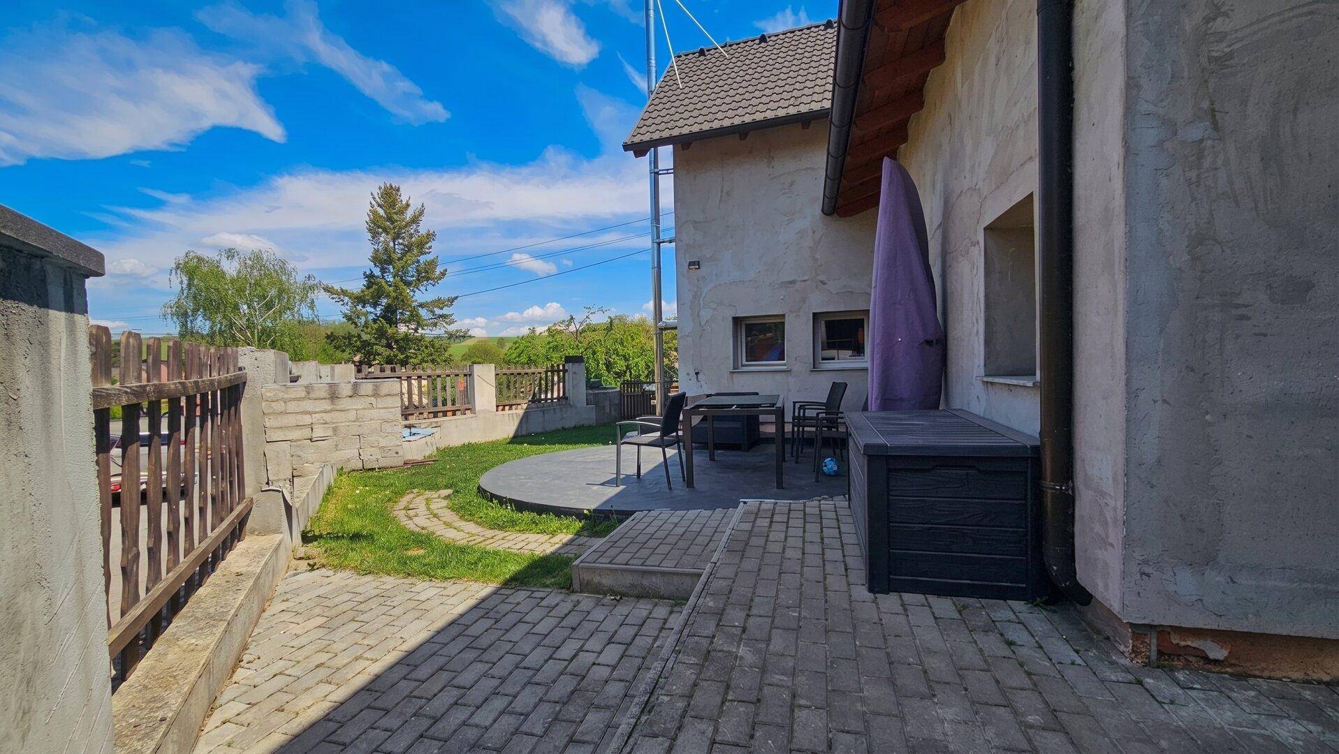 Außenbereich mit gepflasterter Terrasse, runder Betonfläche und Gartenmöbeln neben dem Haus.