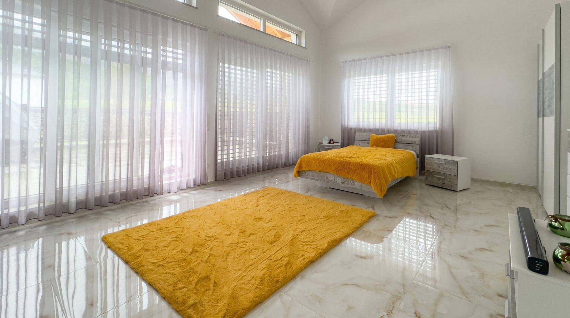 Helles Schlafzimmer mit großen Fenstern, gelbem Teppich und Bettdecke auf Marmorboden.
