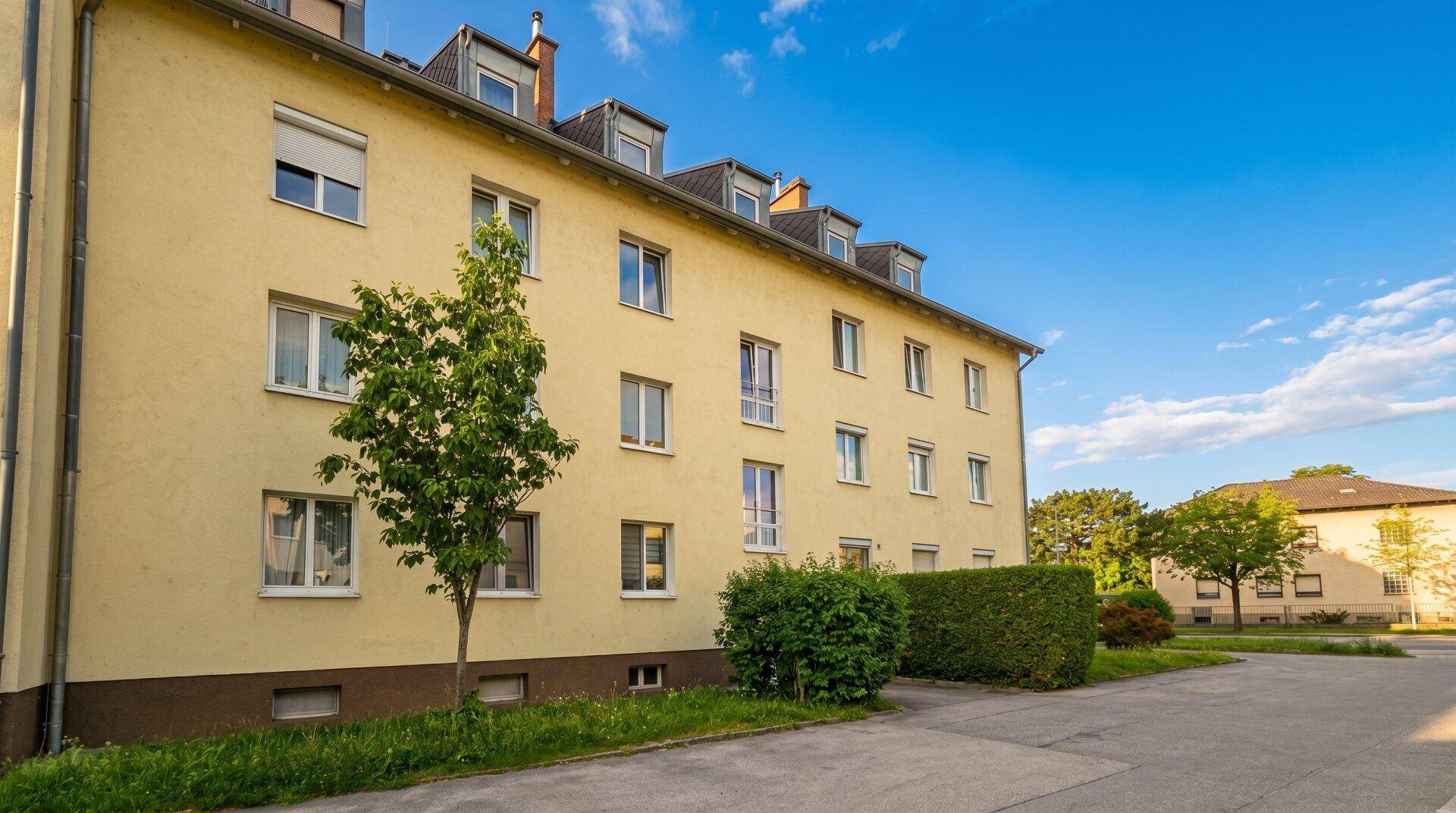 Gelbes Mehrfamilienhaus mit mehreren Fenstern und Dachgauben unter blauem Himmel.