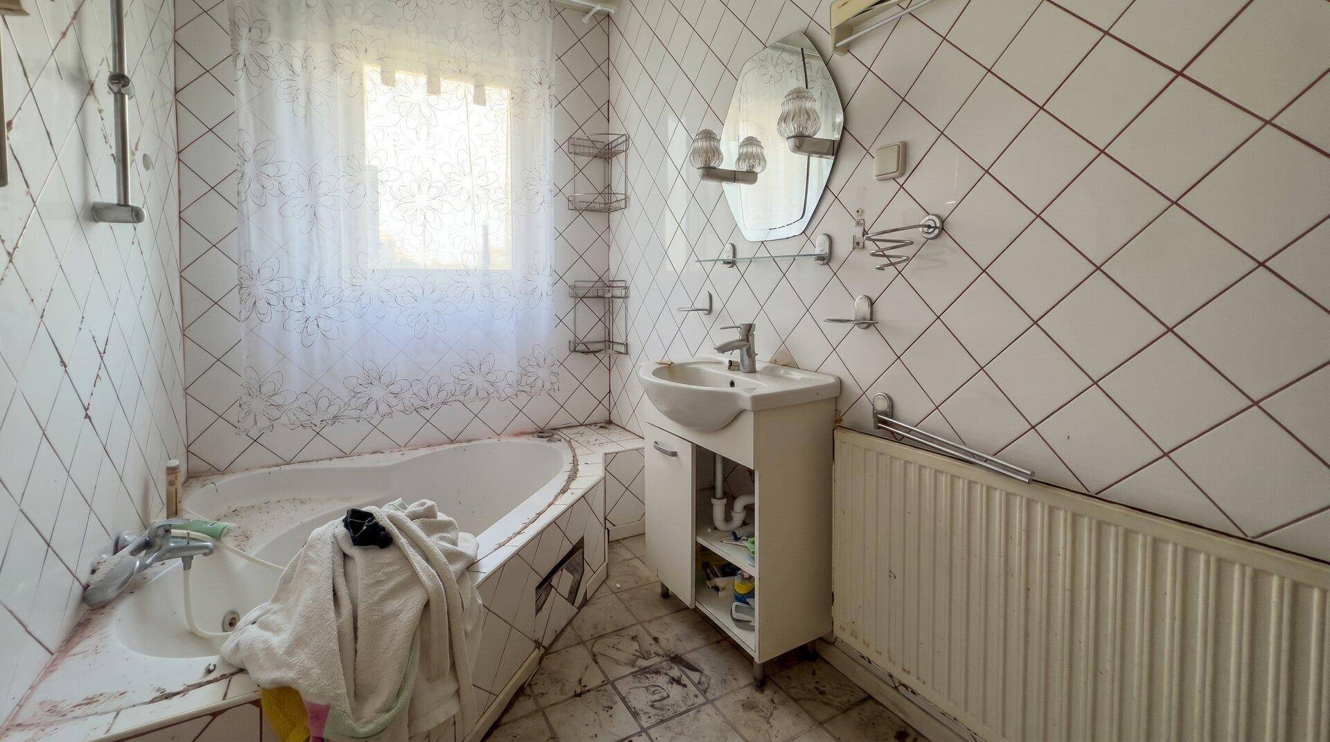 Veraltetes Badezimmer mit Fliesen, Badewanne und Fenster, das eine umfassende Renovierung benötigt.