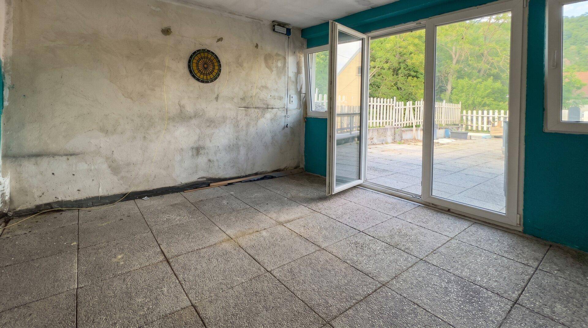 Renovierungsbedürftiger Raum mit Fliesenboden und großen Glastüren, die zur Terrasse führen.