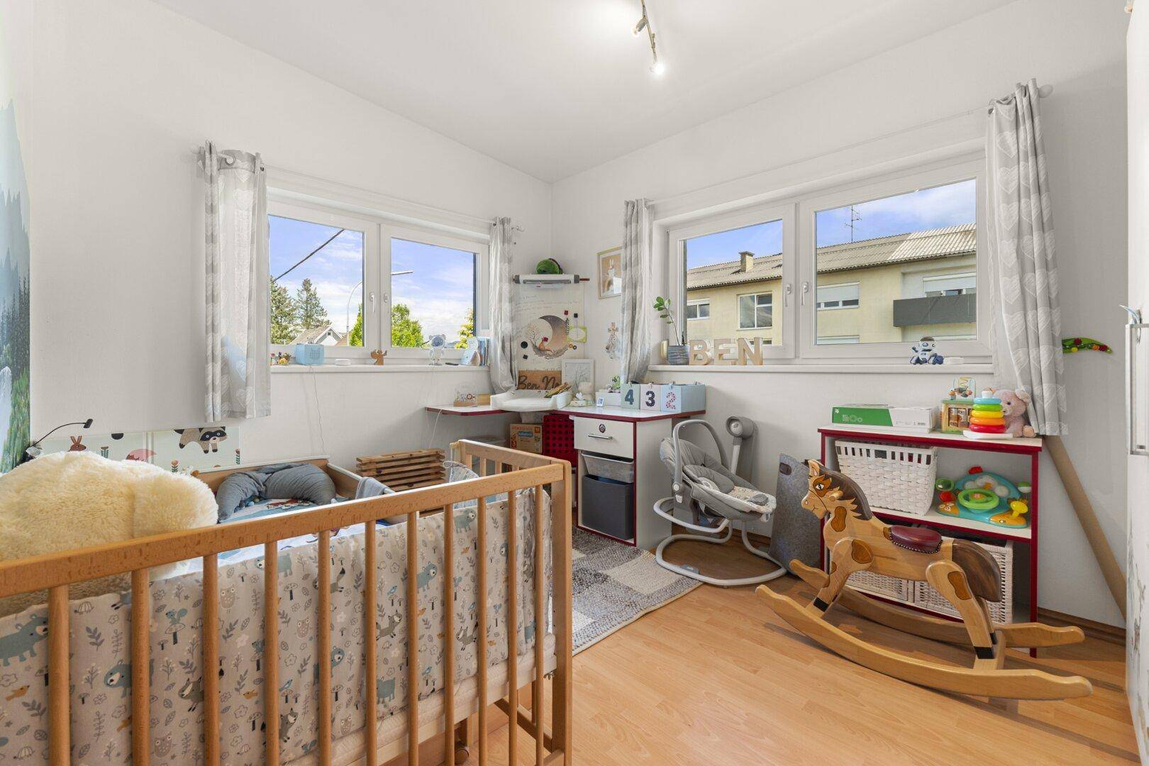 Helles Kinderzimmer mit Babybett, Schaukelpferd und zwei Fenstern für viel Tageslicht.