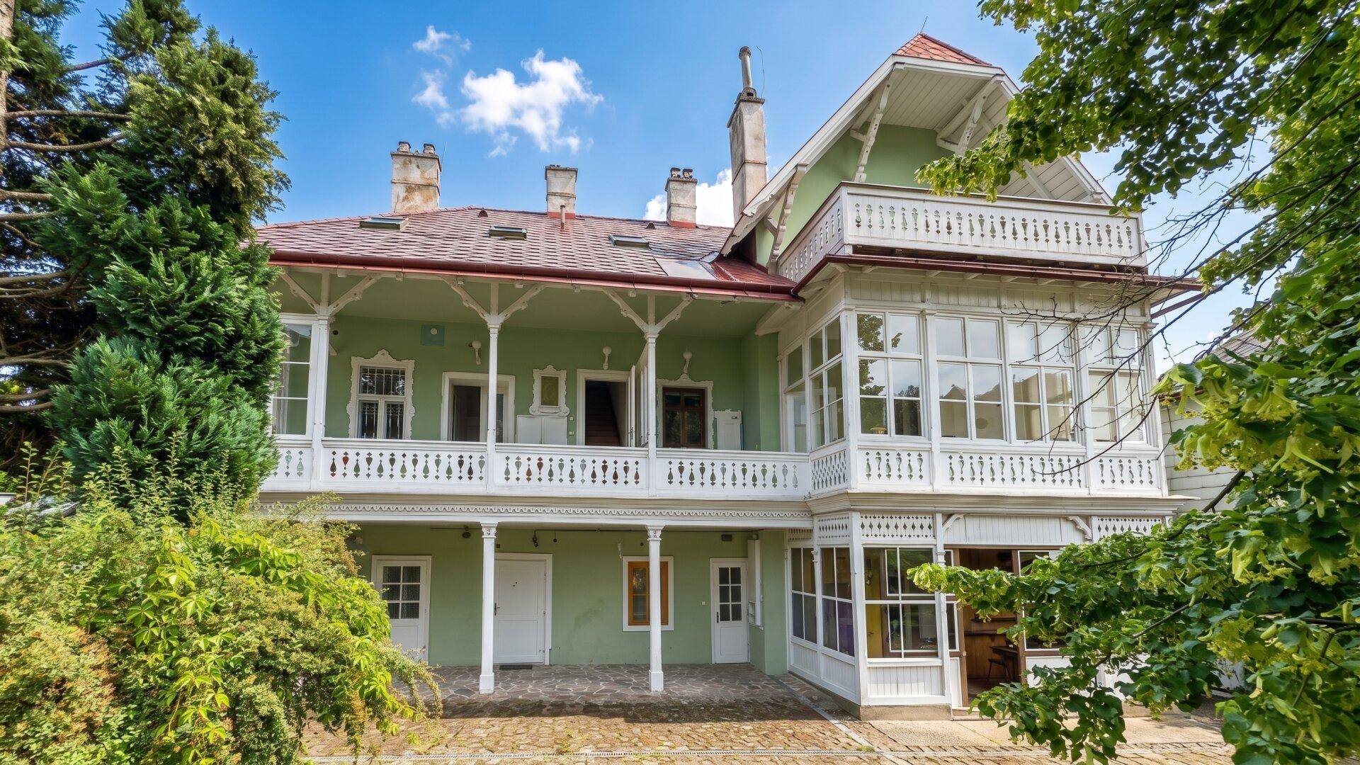 Historisches Gebäude mit grüner Fassade, vielen Balkonen und umgebenden Bäumen im Sonnenlicht.