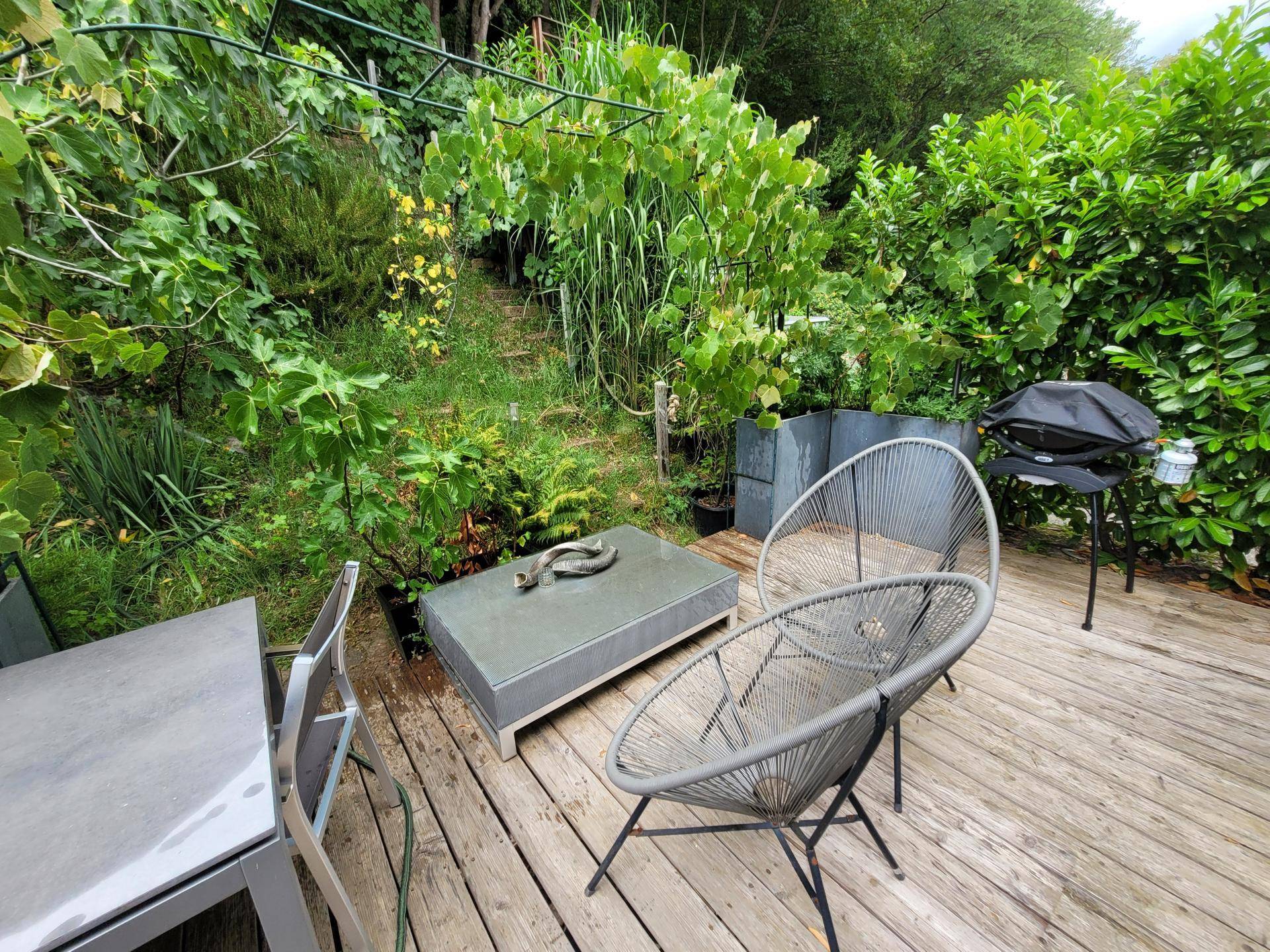 Gemütliche Holzterrasse mit Sitzgelegenheiten und Grillbereich, umgeben von üppigem Grün und Pflanzen.