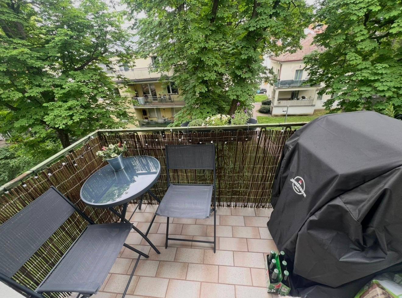 Kleiner Balkon mit Sitzgelegenheiten und Grill, umgeben von grünen Bäumen.