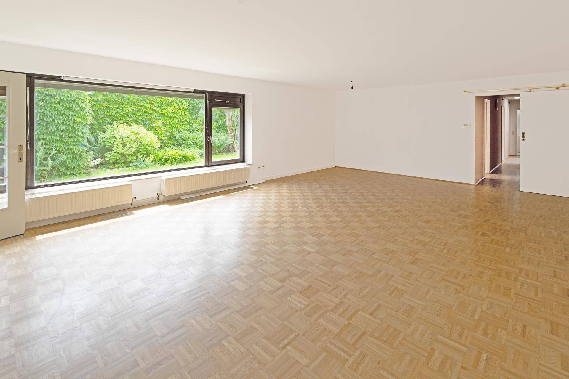 Geräumiges Wohnzimmer mit klassischem Parkettboden und großen Fenstern mit Blick ins Grüne.