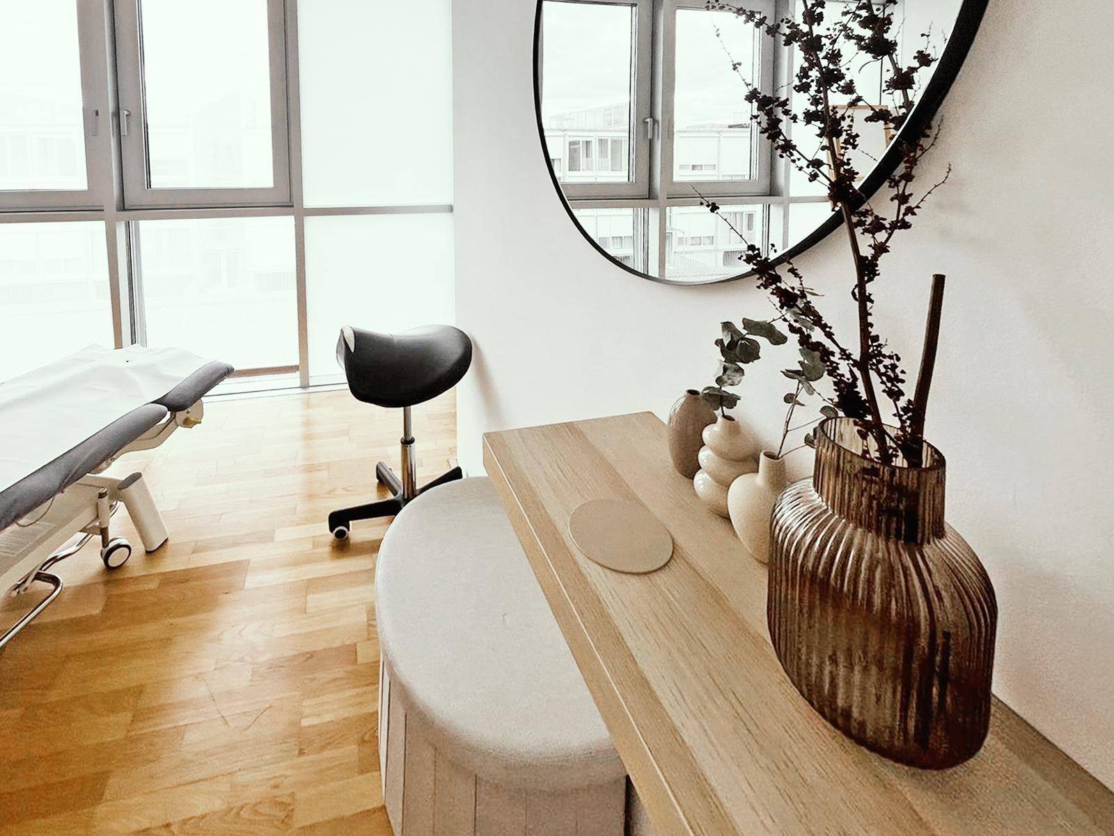 Heller Behandlungsraum mit großen Fenstern, Holzboden und einer stilvollen Holzkonsole mit Dekoration.