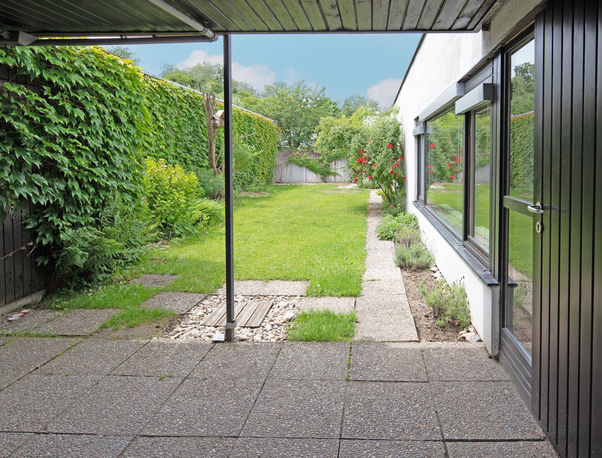 Überdachte Terrasse mit Steinplatten, die in einen grünen Garten mit Rasenfläche übergeht.