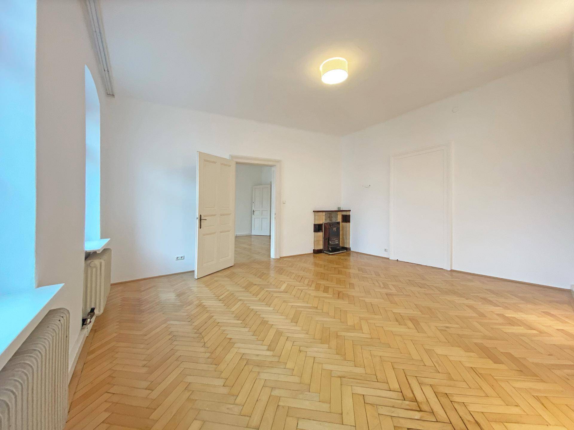 Leeres Zimmer mit Fischgrätparkettboden, weißen Wänden und einem charmanten Kachelofen.