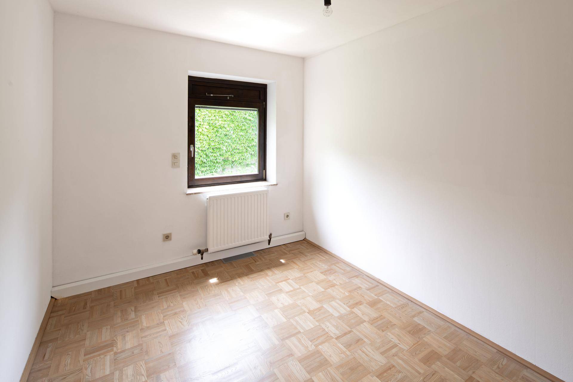 Helles Schlafzimmer mit Parkettboden und einem Fenster, das einen angenehmen Blick ins Grüne bietet.