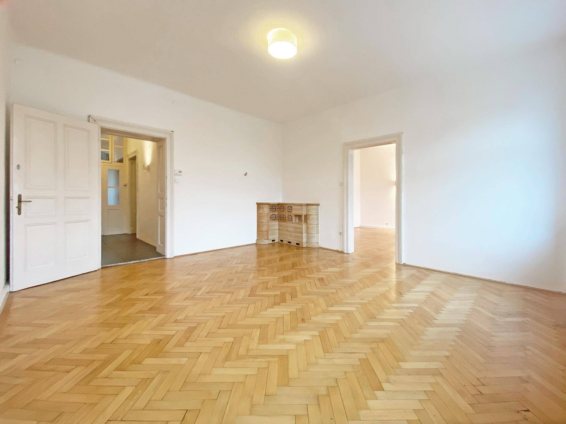 Geräumiges, leeres Zimmer mit klassischem Fischgrätparkett und einem traditionellen Kachelofen.