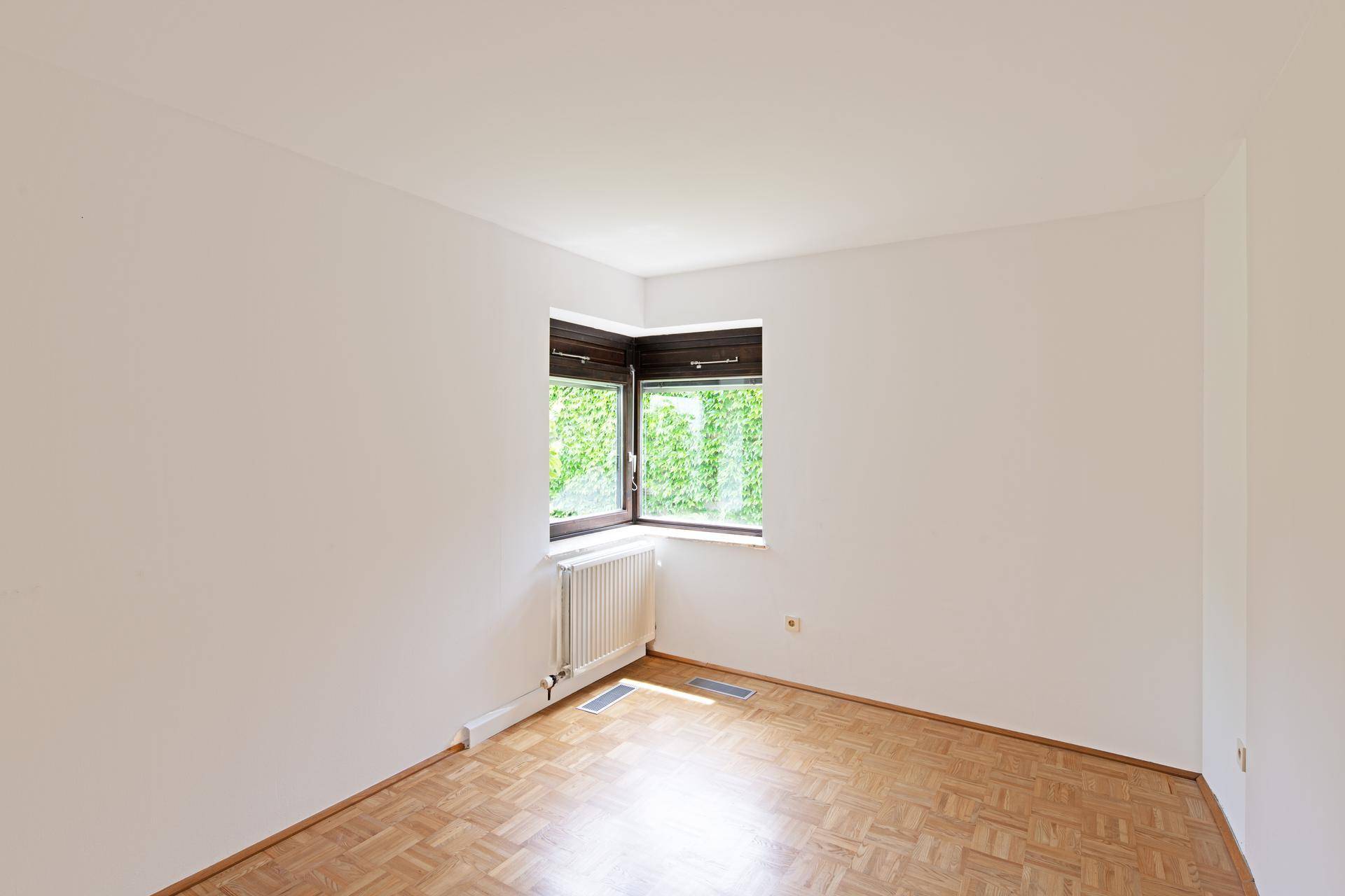 Eckzimmer mit Parkettboden und einem Eckfenster, das eine helle Atmosphäre und Blick ins Grüne schafft.