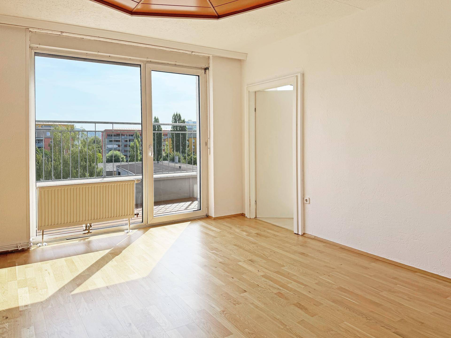 Geräumiger, heller Raum mit Holzboden und großem Fenster mit Zugang zum Balkon.